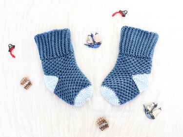 Nautical-inspired pair of blue crochet baby socks with ribbed cuffs and pale-blue toes, photographed with small ship buttons