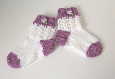 Weiß-lila gestrickte Babysocken mit Spitzenmuster und kleinen lila Rosen am Bündchen auf hellem Hintergrund.