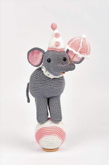 Gehäkelter grauer Amigurumi‑Elefant mit rosa Details auf gestreiftem Ball, hält kleinen Schirm