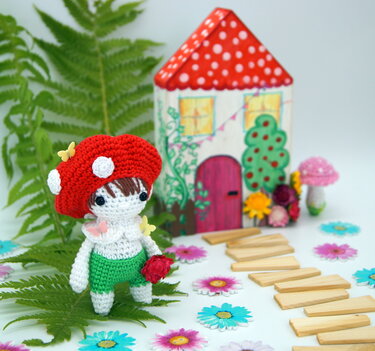 Gehäkelte Amigurumi-Puppe mit rotem Pilzhut, grünen Shorts und roter Blume vor bemaltem Miniaturhaus