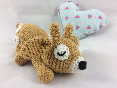 Gehäkeltes liegendes Rehkitz-Amigurumi aus braunem Garn vor blau-rosa Herzkissen