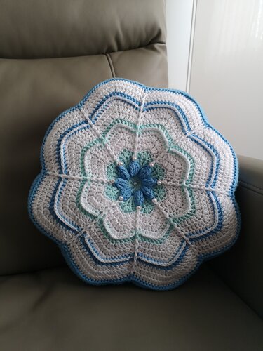 Rundes, blumenförmiges Häkelkissen in Weiß, Hellblau und Türkis mit Perlen, liegend auf einem grauen Sessel.