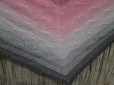 Gestricktes Dreieckstuch in Rosa bis Grau mit wellenförmigem Lochmuster und zentraler Mittelreihe, vor Bambusmatte drapiert