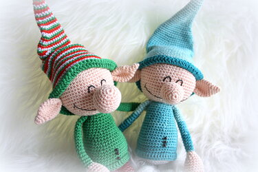 Zwei gehäkelte Amigurumi-Wichtel, links grün mit gestreifter Mütze, rechts türkisblau, beide mit großen Nasen und lächelnden Gesichtern auf weißem Fell