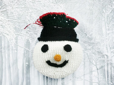 Gehäkelter Schneemannkopf mit schwarzem Zylinder, schwarzen Augen und oranger Nase vor verschneitem Wald.