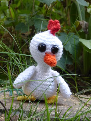 Weißes gehäkeltes Huhn-Amigurumi mit großen schwarzen Augen, rotem Kamm und orangefarbenem Schnabel vor grünem Hintergrund.