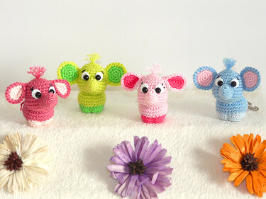 Vier gehäkelte Elefanten-Amigurumi in Pink, Grün, Hellrosa und Blau mit großen runden Augen auf weißem Untergrund. Rechts ist ein Metallring sichtbar, im Vordergrund liegen getrocknete Blumen.