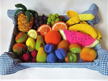 Bunter Korb gefüllt mit vielfältigem gehäkeltem Obst und Früchten wie Ananas, Bananen, Wassermelone und Beeren auf blau-weiß kariertem Tuch.