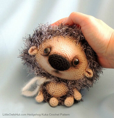 Crocheted hedgehog amigurumi with fuzzy gray spines, beige body, black nose, and brown eyes held by a hand.