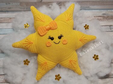 Gelbes gehäkeltes Sternkissen mit Gesicht und Schleife auf weißem Deko‑Vlies