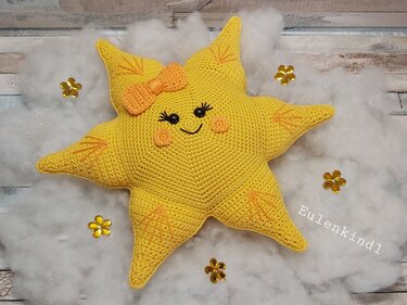 Gehäkeltes gelbes Sternkissen mit lächelndem Gesicht und orangefarbener Schleife auf flauschigem Untergrund