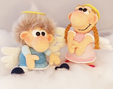 Two crocheted amigurumi angels — one in blue with fuzzy hair and a halo, one in pink with braided hair and layered wings — sitting on a white fluffy surface