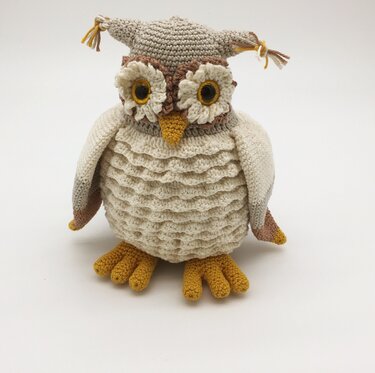 Gehäkelte Amigurumi-Eule in Creme, Beige und Gelb mit großer Augenpartie und Zipfelmütze auf hellem Hintergrund.