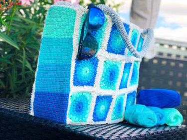 Gehäkelte Henkeltasche in Blau- und Türkistönen mit Granny-Squares, Garnknäueln und Sonnenbrille.