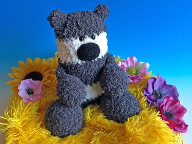 Gehäkelter grauer Amigurumi-Teddy mit weißer Schnauze und schwarzer Nase, sitzend in gelbem Fellnest mit bunten Blumen vor blauem Hintergrund