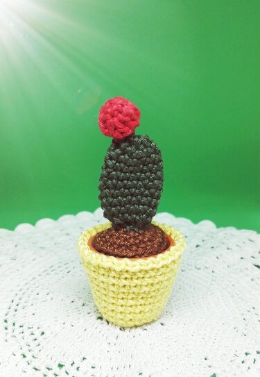 Gehäkelter Mini-Kaktus mit roter Blüte im gelben Topf auf weißem Häkeldeckchen