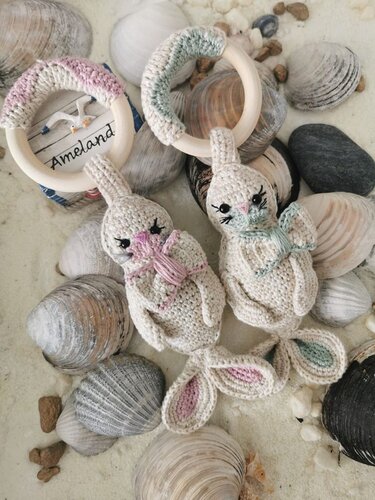 Zwei gehäkelte Hasen-Greiflinge mit Holzringen in Rosa und Mintgrün auf Muscheln und Sand.