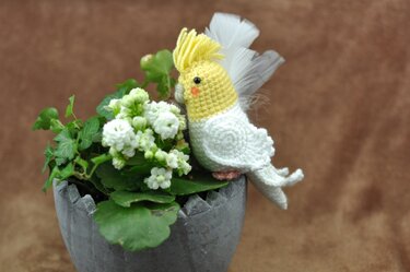 Yellow-crested white crochet cockatiel amigurumi with feather accents perched on a grey planter beside white flowers.