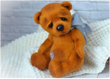 Gehäkelter brauner Teddybär mit hellblauer Schleife und Zierflicken sitzt auf weißer Decke