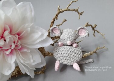 Grau‑rosa gehäkelte Amigurumi‑Maus auf goldenen Zweigen mit großem weißen Blütenkopf im Hintergrund.