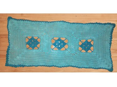 Türkisfarbener gehäkelter Tischläufer mit drei offenen Quadratmotiven und dunklem Rand auf Holztisch.