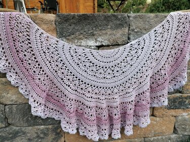 Crescent crochet shawl with lace stitches and scalloped edge in pink gradient draped over a stone wall