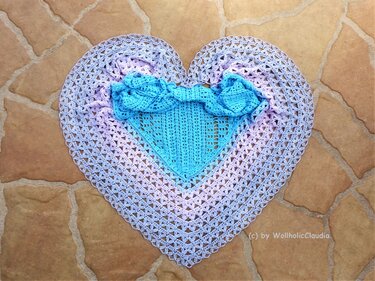Herzförmiges gehäkeltes Tuch in Türkis und Lavendel auf braunem Fliesenboden.