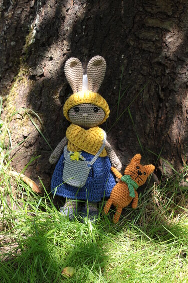 Gehäkelte Hasenpuppe mit Hut, Schal und Kleid neben einer kleinen gehäkelten Katze vor einem Baum