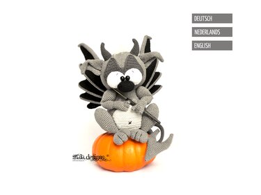 Gehäkelter grau-schwarzer Gargoyle mit Flügeln und Hörnern, sitzend auf einem orangefarbenen Kürbis und mit Häkelnadel in den Händen.