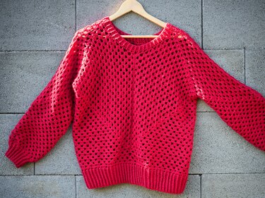 Roter gehäkelter Damenpullover mit offenem Lochmuster an einem Holzbügel vor einer Betonwand