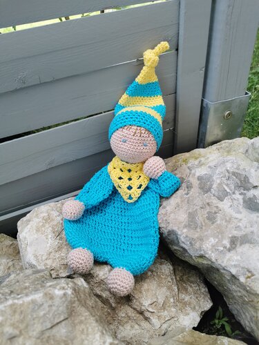 Handgehäkelte Babypuppe in Blau und Gelb mit gestreifter Zipfelmütze sitzt auf Steinen vor einem Holzzaun.