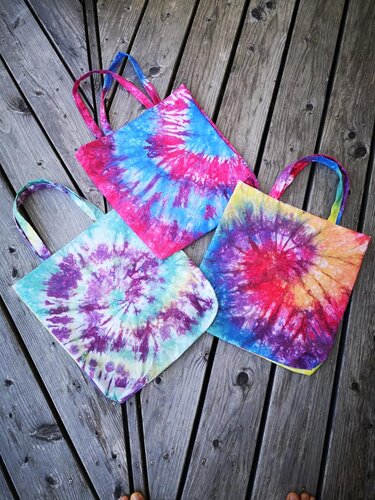 Drei bunte Tie‑Dye Stofftaschen mit Spiralmustern auf Holzdielen.