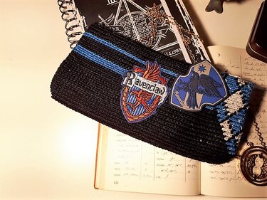 Schwarze gehäkelte Tasche mit blauen Streifen und Wappen-Patches auf offenem Buch