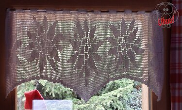 Weißer, gehäkelter Fensterbehang mit filigranen Schneeflockenmotiven vor einem Fenster, im Hintergrund grünes Gehölz