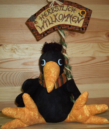 Handgefertigte schwarze Stoffkrähe mit orangefarbenem Schnabel und Beinen vor einem Schild mit der Aufschrift "HERBSTLICH WILLKOMMEN".