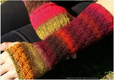 Gestrickte, fingerlose Armstulpen in Rot-, Orange- und Brauntönen mit Streifen- und Rippenmuster, über den Unterarmen gefaltet.