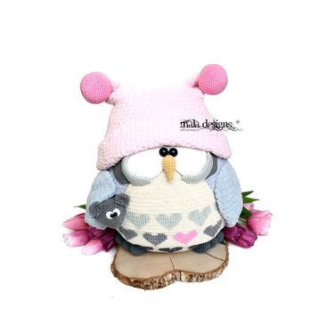 Crocheted amigurumi owl wearing a pink bobble hat, heart-patterned belly, holding a small grey amigurumi