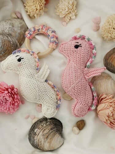 Zwei gehäkelte Seepferdchen (weiß und rosa) und ein Greifring aus Holz mit Häkelbezug auf hellem Stoff, umgeben von Muscheln und Trockenblumen.