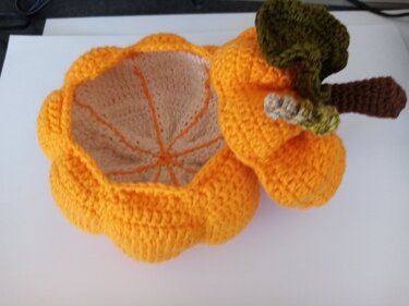 Orange crocheted pumpkin-shaped storage basket with lid, brown stem and green leaf, lid open to show beige interior.