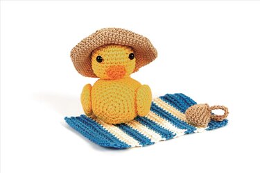 Gehäkeltes gelbes Entchen mit braunem Strohhut auf blau-weiß gestreiftem Strandtuch, neben einer kleinen gehäkelten Tasse