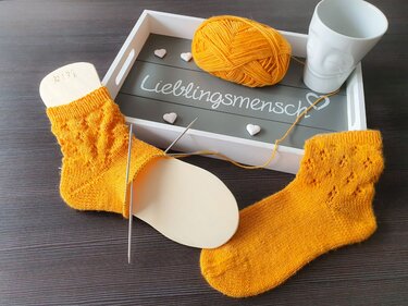 Gelbe gestrickte Kindersocken, eine auf Sockenbrett gespannt, daneben eine fertige Socke und Wollknäuel