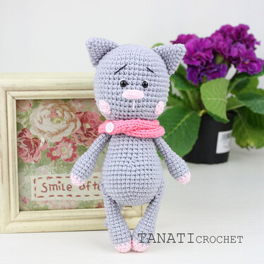 Gehäkelte graue Amigurumi-Katze mit rosa Schal vor Blumenbild