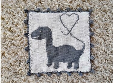 Gestricktes Quadrat in Weiß und Grau mit Dinosaurier- und Herzmotiv und Bommelrand