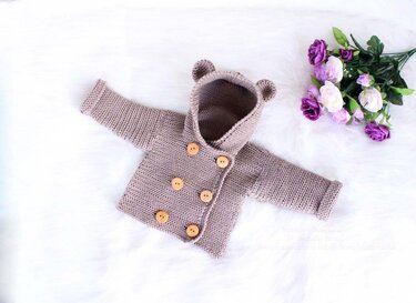 Hellbraune gehäkelte Babyjacke mit Kapuze und Bärenohren, mit sechs Holzknöpfen, auf weißem Fellstoff neben violetten Blumen.