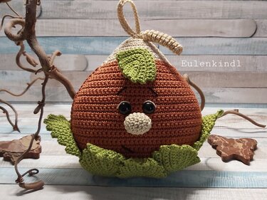 Gehäkelte braune Eichel-Amigurumi mit grünen Blättern, beigefarbener Kappe und Gesicht auf hellem Holzhintergrund.