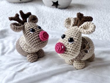 Two amigurumi reindeer, one light beige with a star button, the other brown, both with red noses, on a white textured surface.
