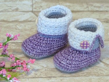 Zwei lavendelfarbene, gehäkelte Babyschuhe mit weißem Umschlag und kariertem Knopf neben kleinen rosa Blüten