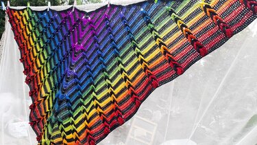 Gehäkeltes Dreieckstuch mit Zickzackmuster in Regenbogenfarben, an einer Wäscheleine im Freien aufgehängt.
