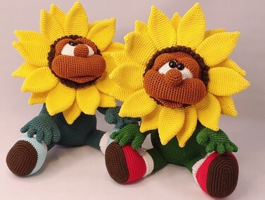 Two crocheted amigurumi sunflowers with layered yellow petals, brown faces, green bodies and striped feet sitting side by side.