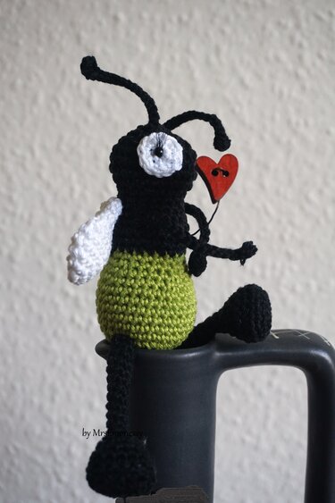 Gehäkeltes Amigurumi Glühwürmchen in Schwarz und Grün mit rotem Herz auf Tassenrand